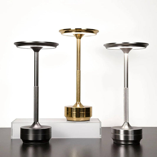 Three Lumin Lamps in black, gold, and silver are showcased on a plain background. These rechargeable LED lamps feature an elegant design with a modern cylindrical base and a circular dish-like top. The gold lamp is set on a transparent rectangular block for added impact.